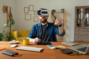 Wall Mural - Serious bearded designer sitting at table and drawing on digitizer while using virtual reality simulator at home