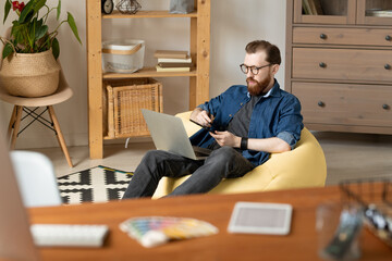 Wall Mural - Confident young layout artist with beard sitting in yellow bean bag and using digitizer tablet while working from home