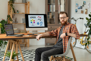 Wall Mural - Portrait of serious handsome bearded brand designer sitting at desk with desktop computer and laptop in home office