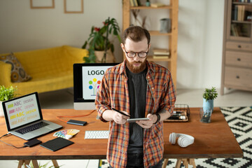 Wall Mural - Serious hipster bearded designer in eyeglasses using stylus while checking sketch on tablet in cozy home office