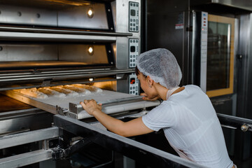 Wall Mural - Young caucasian woman baker is putting the bread dough and in electric oven at baking manufacture factory.