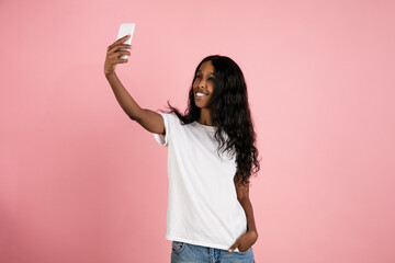 Wall Mural - Taking selfie. Cheerful african-american young woman isolated on pink background, emotional and expressive. Concept of human emotions, facial expression, sales, ad. Beautiful model with long curly