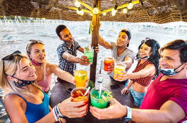 Wall Mural - Young trendy friends toasting at beach cocktail bar chiringuito with face mask - New normal summer concept with people having fun together cheering drinks - Warm dark filter with focus on central guys