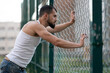 © Vladimir - Male athlete stands on the Playground and looks through a gap in the fence. A man looks into the distance in the city