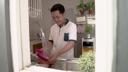 Wall Mural - Happy young asian man in white shirt standing and washing dishes on the kitchen