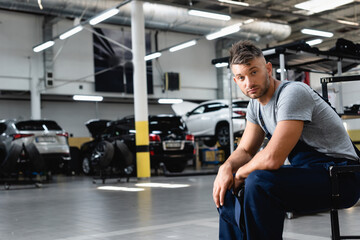 Wall Mural - tired technician in overalls holding cap and looking at camera while sitting near cars in workshop