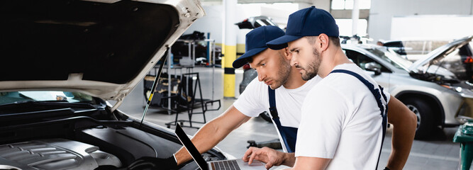 Wall Mural - Panoramic shot of auto mechanics looking at laptop screen near car at service station