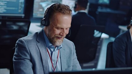 Joyful Bearded Senior Technical Support Specialist is Talking on a Headset while Working on a Computer in a Dark Monitoring and Control Room Filled with Computer Display Screens and Data Servers.