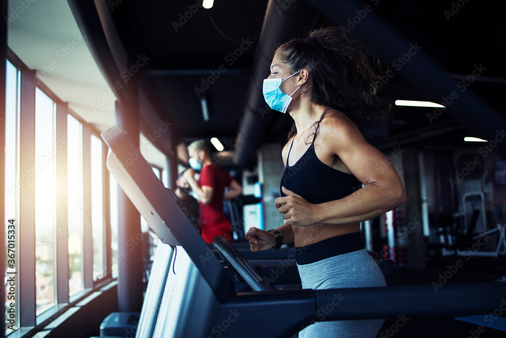 Active fitness woman wearing hygienic protective face mask while training in gym to protect herself and others against coronavirus or covid-19. Stay safe and healthy. - obrazy, fototapety, plakaty 