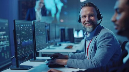 Bearded Happy Senior Technical Support Specialist is Talking on a Headset while Working on a Computer. Successful Employee Turns to the Camera and Gently Smiles. Monitoring Room with Display Screens.