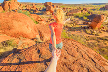 Follow Me, Tourist Woman Holding Hands At Top Of Karlu Karlu. Aerial View Of Vibrant Colors Of Natural Boulders Rocks At Devils Marbles, Northern Territory, Australia. Concept Hand In Hand Couple.