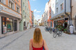 Looking over shoulder of young blond woman in red dress with big red shopping bag standing in the middle of historic shopping pedestrian zone with old houses on sunny summer day