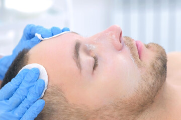 Portrait of man on cleaning face procedure by woman cosmetologist in beauty clinic, side view. Beutician doctor wiping patient's face with cotton pads. Beauty industry concept. Guy is lying on couch.