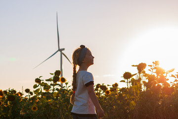 Cute girl in white t-shirt smelling sunflower in sunset field with wind turbines farm on background. Child with long braid hair on countryside landscape with yellow flowers. Farming concept wallpaper.