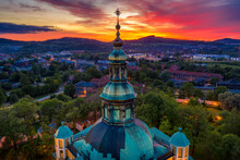 Aerial View Of Beautifully Decorated Tower On The Roof Of A Baroque Church In Jelenia Gora. Temple Surrounded By Karkonosze Mountains At Red Sunset Sky. View Of The City Skyline From The Drone
