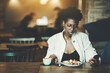 © skyNext - A portrait with a copy space place on the left of a young dainty black woman in elegant spectacles and white trench having her lunch alone in a cozy restaurant nook and using a smartphone