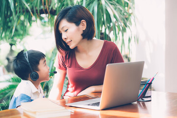 Wall Mural - Young asian mother and son using laptop computer for study and learning together at home, boy wearing headphone for e-learning with distancing, teacher or mom support child, education concept.