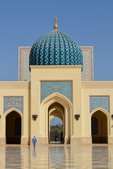 A man walking alone in the inner courtyard of Sultan Qaboos Mosque. On the background, there are three passages with arches and a beautiful blue dome. Sohar, Oman.