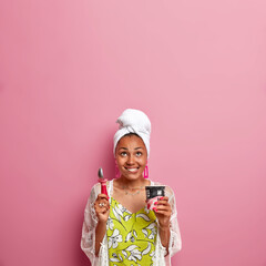 Wall Mural - Vertical shot of happy lady with dark skin, bites lips and looks above, eats delicious frozen dessert, holds spoon, enjoys strawberry ice cream, wears wrapped towel, casual dress, empty space upwards