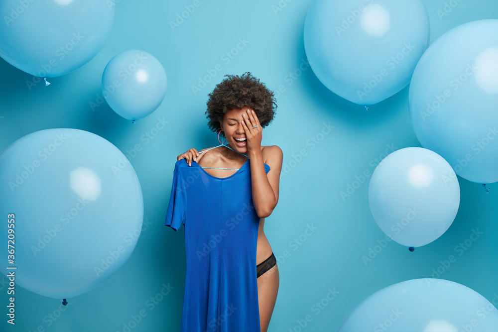 Joyful undressed Afro American young woman holds dress on hangers, dresses for blue theme wedding celebration, laughs positively, poses against blue background, big balloons. Monochrome shot - obrazy, fototapety, plakaty 