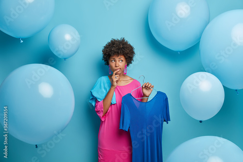 Indecisive thoughtful African American woman chooses outfit for banquet, thinks what to wear, holds dress on hangers, isolated over blue background with big balloons. Women, fashion, clothing