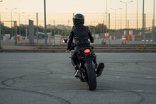 Back View Of Stylish Woman In Black Leather Jacket, Pants And Protective Helmet Rides On Sports Motorcycle At Urban Outdoors Parking.