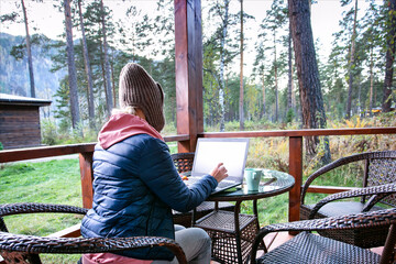 Female blogger concentrated on keyboarding on netbook article for web page sitting at table on terrace of house in autumn forest.