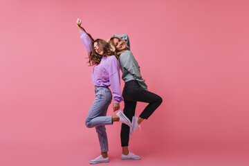 Wall Mural - Full-length photo of inspired girls funny dancing together. Indoor shot of cheerful best friends standing on bright background.