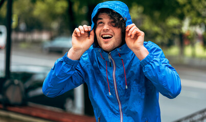 Wall Mural - Image of man smiling broadly, wearing blue raincoat during the rain outside. Handsome happy male in blue raincoat enjoying the rain in the city street. The guy has joyful expression in rainy weather.