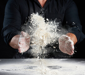 Wall Mural - chef in black uniform sprinkles white wheat flour in different directions, product scatters dust