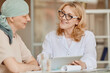 Warm-toned portrait of smiling female doctor talking to bald woman and showing data at digital tablet during consultation on alopecia and cancer recovery, copy space