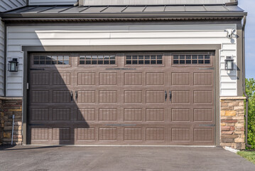 Double car classic insulated steel raised panel brown garage door framed with a white trim to add accent, with transom light windows divided by muntins on a new American home