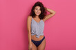 Adorable dark haired woman wearing t-shirt and panties posing against pink background. Pretty beautiful girl with curly hair keeping hand on head.