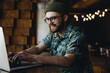 © mykolastock - Smiling young smart bearded male hipster in eyeglasses and hat having video meeting on laptop in online communications platform