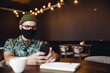 © mykolastock - Fashionable smart hipster male wearing handmade face mask  using smartphone in cafe