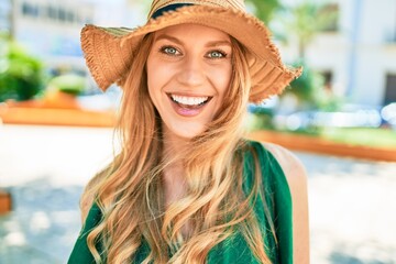 Young beautiful blonde woman on vacation wearing summer hat smiling happy. Standing with smile on face at street of city.