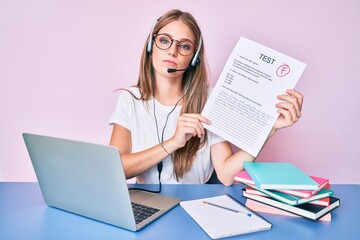 Young blonde girl showing failed exam sitting on the table thinking attitude and sober expression looking self confident