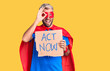Young blond man wearing super hero costume holding act now cardboard banner smiling happy doing ok sign with hand on eye looking through fingers