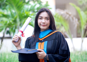 Portrait of happy and excited of young Asian female university graduate wears graduation gown and hat celebrates with degree in university campus in the commencement day. Education concept.