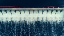 Aerial Flyover Shot Of A Dam In Full Release Mode. The Release Creates This White Ink Like Texture On The Surface Of The Dark Blue Water.