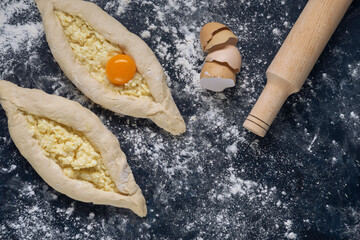 Two raw khachapuri with egg yolk ready for baking on blue textured backdrop. Process of making bakery. Adjarian Khachapuri Recipe – Georgian cheese bread. Top view. Flat lay. Space for text.