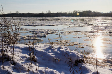 Ice drift on a river with blue high water and big water, white snow broken ice full of hummocks in it and tree branches in the foreground in sunny spring day.