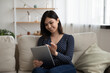 © fizkes - Smiling Asian millennial girl sit relax on couch in living room browse internet on modern tablet gadget, happy Vietnamese young woman rest on sofa at home using pad device, technology concept