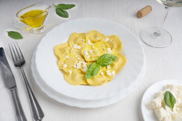 Wall Mural - Italian ravioli pasta in a round plate decorated by fresh basil leaves surrounded by olive oil gravy boat, knife and fork and glass of wine ready for eating on white wooden background at restaurant