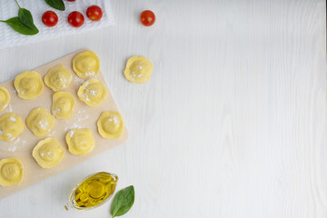Wall Mural - Top view of raw italian ravioli pasta on cutting board surrounded by olive oil gravy boat, spinach and tomato on towel on white wooden background. Image with copy space, horizontal orientation