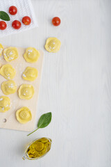 Wall Mural - Top view of uncooked italian ravioli pasta on cutting board surrounded by olive oil gravy boat, spinach and tomatoes on towel on white wooden background. Image with copy space, vertical orientation