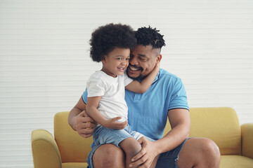 An African American little son sits on his father's lap on sofa at home. Young dad with beard hug and smile at his boy