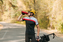 Portrait Of A Thirsty Cyclist In A Sports Outfit Standing On The Road Outside In The Fall, Drinking Water From A Red Bottle And Resting.Tired Cyclist Is Resting, Quenching His Thirst For Water.