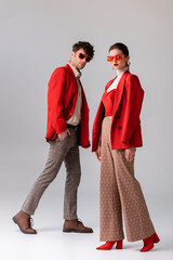 Wall Mural - full length view of fashionable couple in red blazers and sunglasses looking at camera on grey