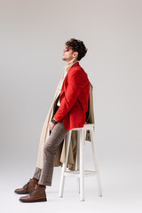 Wall Mural - side view of young man in trendy autumn outfit looking away while leaning on stool on grey
